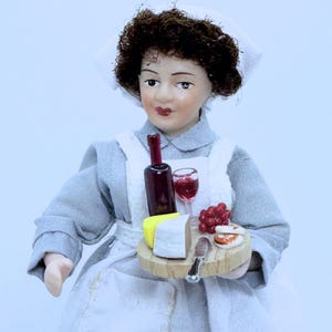 Könnte beinhalten: Eine Porzellanpuppe, die als Dienstmädchen verkleidet ist, hält ein Holztablett mit einer Weinflasche, einem Weinglas, Käse, Trauben und Crackern.
