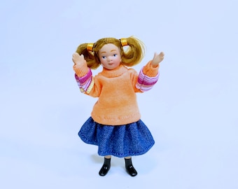 Puede incluir: Una muñeca de porcelana con el pelo rubio en coletas que lleva un jersey de color melocotón y una falda vaquera azul.