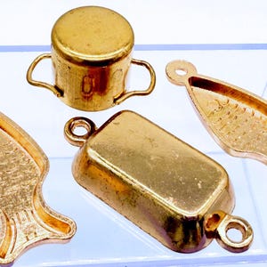 Könnte beinhalten: Eine Sammlung kleiner, goldfarbener Metallanhänger. Die Anhänger umfassen einen kleinen Topf mit Griffen, zwei fischförmige Anhänger und einen rechteckigen Anhänger. Jeder Anhänger hat eine Schlaufe zum Befestigen.