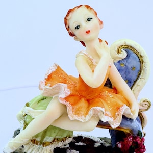 May include: A porcelain figurine of a ballerina wearing an orange and white tutu, sitting on a blue and gold chair. The figurine is detailed with delicate features and a soft, realistic look.