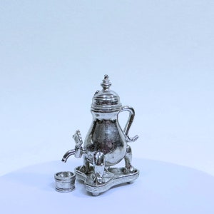 May include: A miniature silver-colored samovar with a spigot, handle, and ornate detailing. A small matching cup sits beside the samovar on a decorative base. The samovar has a domed lid and is likely a decorative item.