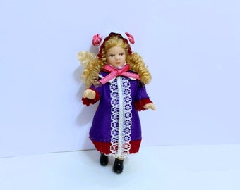 Puede incluir: Una muñeca en miniatura con cabello rubio rizado, vestida con un abrigo morado con ribete de encaje blanco y detalles en rojo. La muñeca tiene un gorro blanco con rosas rosas y un lazo rosa. Zapatos negros completan el atuendo.