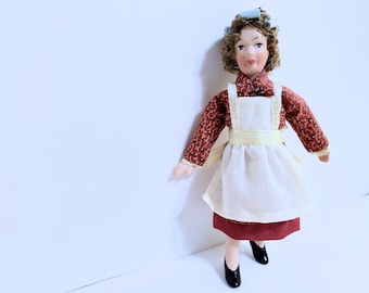 Puede incluir: Una muñeca de porcelana que lleva un vestido rojo y blanco con un delantal blanco y rulos azules. La muñeca tiene una cara pintada con ojos marrones y una tez clara.