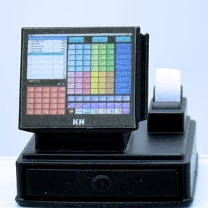 May include: A miniature black cash register with a colorful screen display. The screen shows a grid of colorful blocks and text. A white receipt roll sits on top. The letters "KH" are visible on the screen.