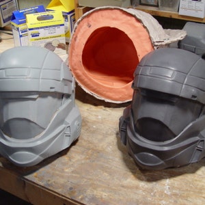 May include: Two grey plastic helmets, one with a darker grey finish, are displayed on a wooden surface. The helmets have a futuristic design with a visor and a textured surface.