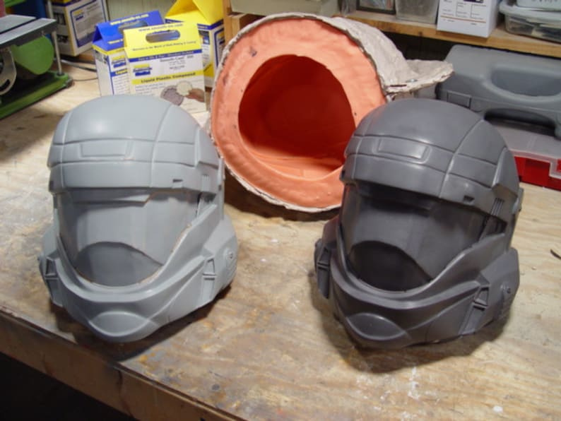 May include: Two grey plastic helmets, one with a darker grey finish, are displayed on a wooden surface. The helmets have a futuristic design with a visor and a textured surface.