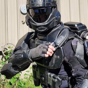 Puede incluir: Un casco negro de estilo futurista con una visera y un traje de armadura negro con una placa de pecho y hombreras.