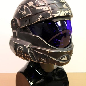 May include: A black helmet with a blue visor and a weathered, distressed finish. The helmet has a white inscription that reads "DDM".