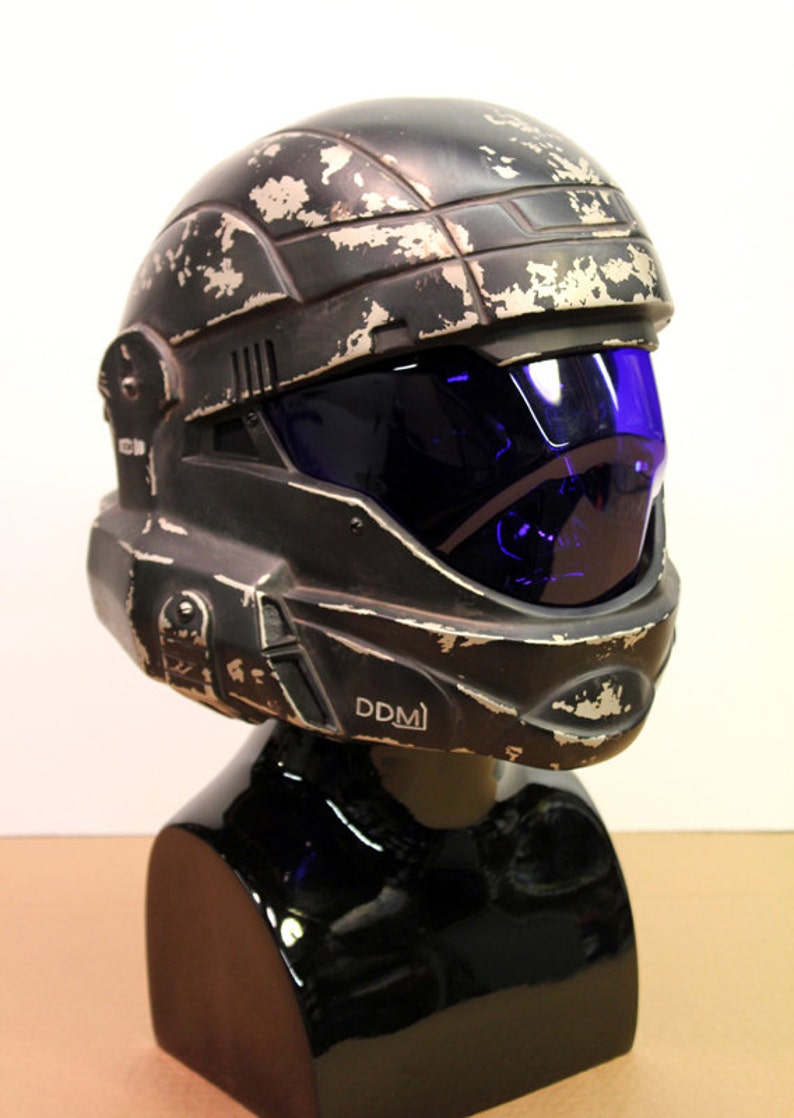 May include: A black helmet with a blue visor and a weathered, distressed finish. The helmet has a white inscription that reads "DDM".