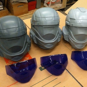 May include: Three grey helmets with blue visors. The helmets have a futuristic design and are made of a smooth, hard plastic. The visors are clear and have a slight tint.