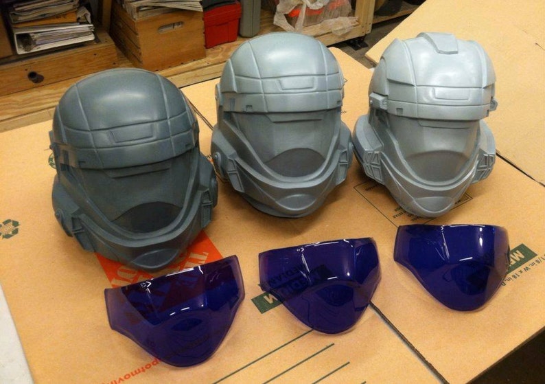 May include: Three grey helmets with blue visors. The helmets have a futuristic design and are made of a smooth, hard plastic. The visors are clear and have a slight tint.
