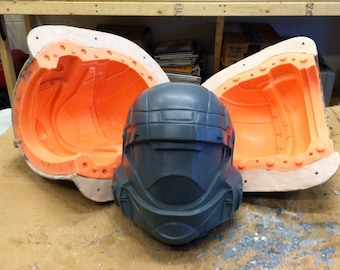 Op de afbeelding: Twee oranje en witte siliconen mallen voor het gieten van een helm, met een donkergrijze helm in het midden.
