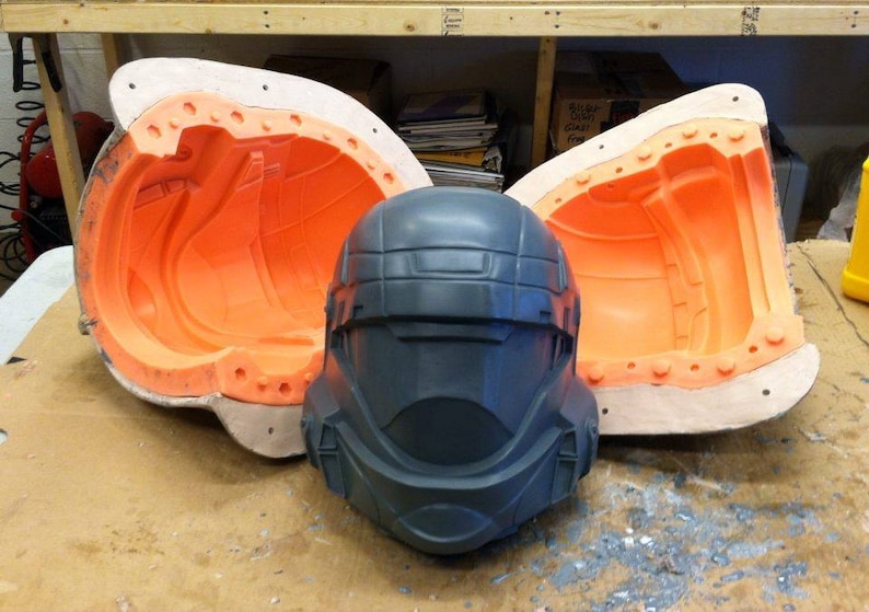 May include: Two orange and white silicone moulds for casting a helmet, with a dark grey helmet in the centre.