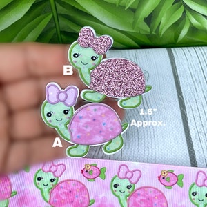 May include: Two turtle-shaped embellishments, approximately 1.5 inches in size, with green bodies and pink shells. One has a glittery shell and a pink bow. The other has a pink shell with a polka dot pattern and a pink bow. A pink ribbon with turtle and fish designs is also visible.