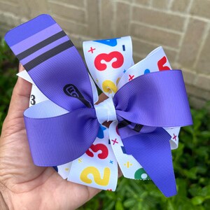 May include: Purple and white hair bow with a colorful number and plus sign pattern. The bow is made of ribbon and has a white center.