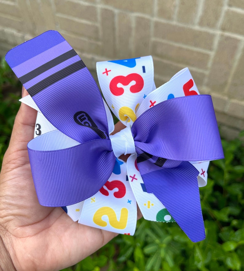 May include: Purple and white hair bow with a colorful number and plus sign pattern. The bow is made of ribbon and has a white center.