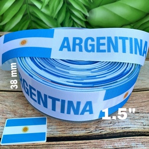May include: A roll of white and blue ribbon with the Argentinian flag and the word "ARGENTINA" printed on it. The ribbon is 1.5 inches wide and 38 mm in diameter.