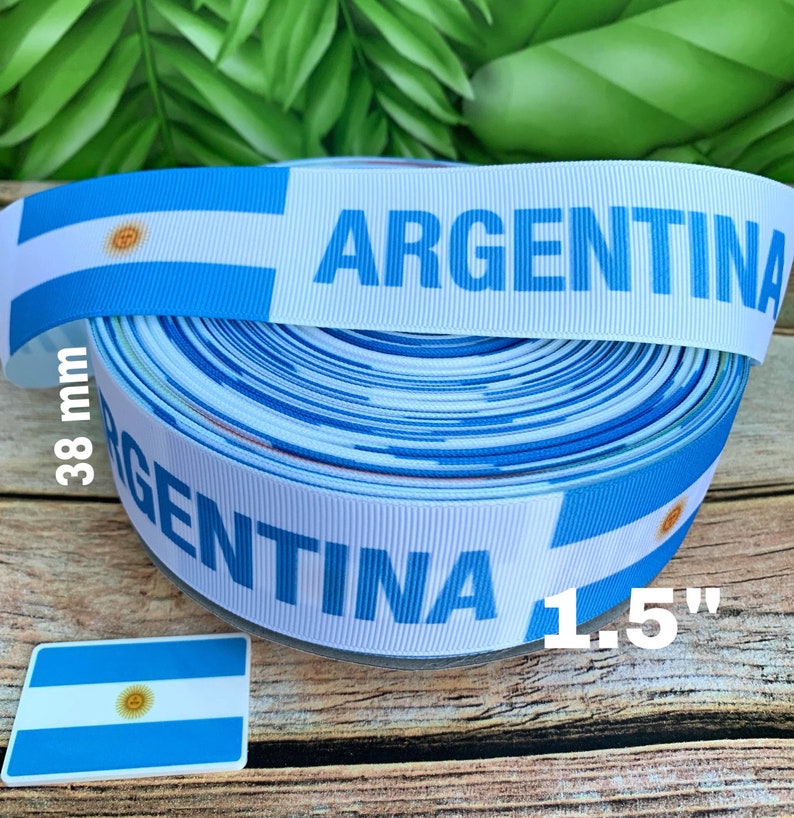 May include: A roll of white and blue ribbon with the Argentinian flag and the word "ARGENTINA" printed on it. The ribbon is 1.5 inches wide and 38 mm in diameter.