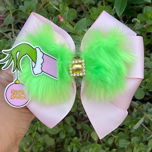 Peut inclure: Un nœud de cheveux de Noël rose et vert avec une main du Grinch tenant un ornement de Noël qui dit "Joyeux Noël".
