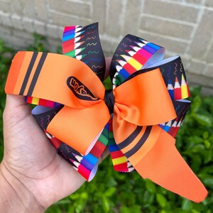 May include: Orange and black crayon-themed hair bow with a Crayola logo. The bow is made of ribbon and has a colorful pencil print.