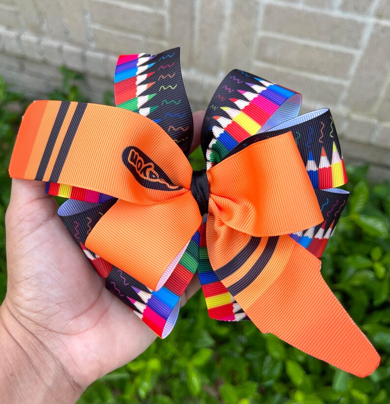 May include: Orange and black crayon-themed hair bow with a Crayola logo. The bow is made of ribbon and has a colorful pencil print.
