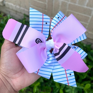 May include: A pink and white bow with a notebook paper design. The bow has black and white stripes and a pink and white striped design. The bow is tied with a white ribbon.