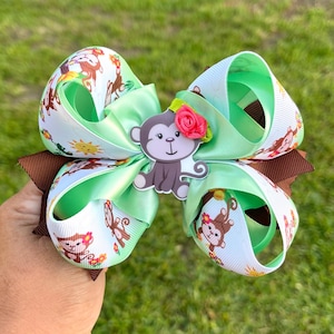 May include: A large, handmade hair bow featuring a monkey design. The bow has layers of light green, brown, and white ribbons. The white ribbon has a repeating pattern of monkeys, flowers, and suns. A monkey embellishment with a pink rose is centered on the bow.
