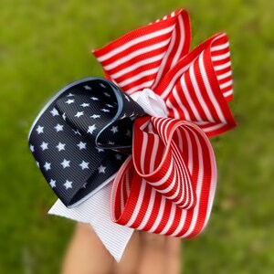 May include: A red, white, and blue ribbon bow with a star pattern. The bow is made of three different ribbons, each with a different pattern. The bow is tied in a knot and is ready to be worn.