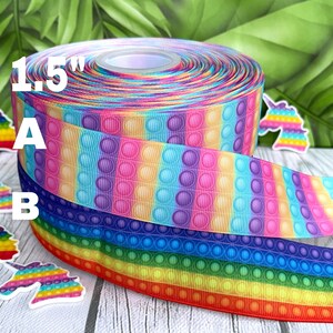 May include: A roll of 1.5 inch wide rainbow pop it bubble ribbon. The ribbon has a repeating pattern of rainbow coloured stripes with purple, blue, yellow, pink, and orange bubbles. The ribbon is wrapped around a spool and there are unicorn stickers on the table.