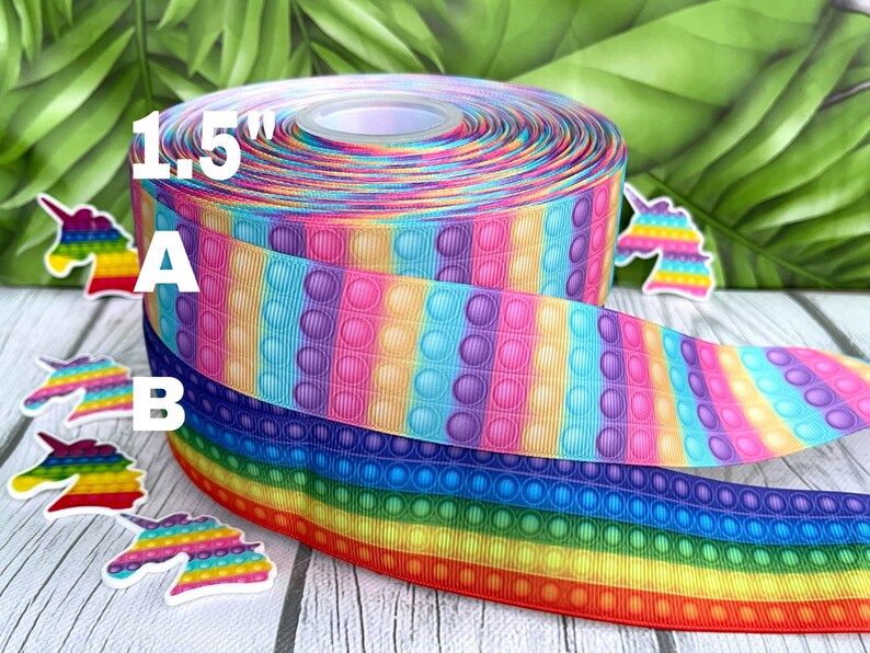 May include: A roll of 1.5 inch wide rainbow pop it bubble ribbon. The ribbon has a repeating pattern of rainbow coloured stripes with purple, blue, yellow, pink, and orange bubbles. The ribbon is wrapped around a spool and there are unicorn stickers on the table.