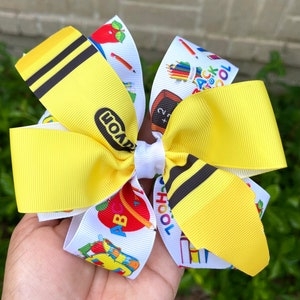 May include: A yellow and white school-themed hair bow with a crayon print and the word "Crayola" on one side and a back to school print with apples, pencils, and the words "Back to School" on the other side.