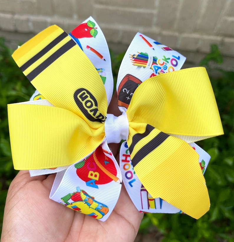 May include: A yellow and white school-themed hair bow with a crayon print and the word "Crayola" on one side and a back to school print with apples, pencils, and the words "Back to School" on the other side.
