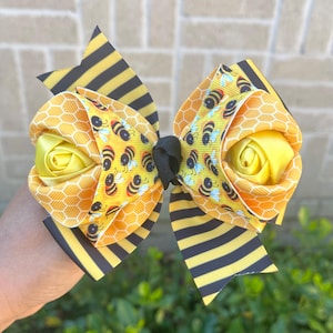 May include: A large hair bow with a bee theme. It features layers of yellow and black striped ribbon, honeycomb patterned ribbon, and a yellow satin rose. The centre is a black knot, and the bow is decorated with bee graphics.