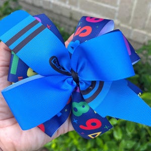 May include: A blue and navy blue hair bow with a colorful number and geometric pattern. The bow is tied with a black ribbon.