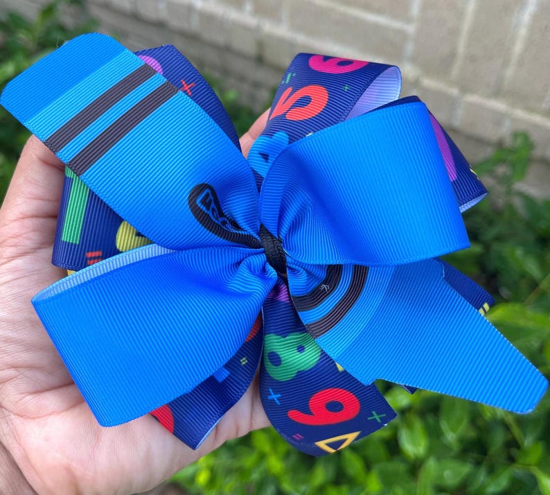 May include: A blue and navy blue hair bow with a colorful number and geometric pattern. The bow is tied with a black ribbon.