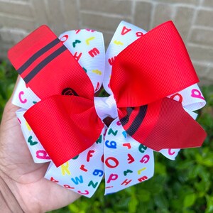 May include: A red and white hair bow with a crayon design and colorful letters on the white ribbon. The bow is tied in a large, fluffy knot.
