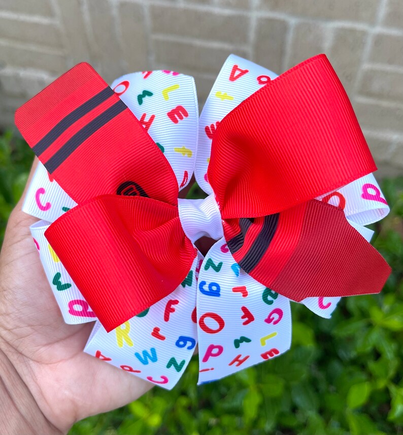 May include: A red and white hair bow with a crayon design and colorful letters on the white ribbon. The bow is tied in a large, fluffy knot.