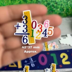 May include: A colorful math problem with a ruler and numbers. The problem is 3036 + 31245 = 6. The numbers are in different colors and the ruler is yellow with black markings. The image is approximately 42 mm by 37 mm.