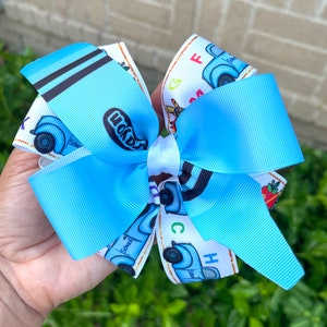 May include: A blue and white hair bow with a school bus design. The bus has the word "School" written on it and the letters "ABC" are printed on the bow.