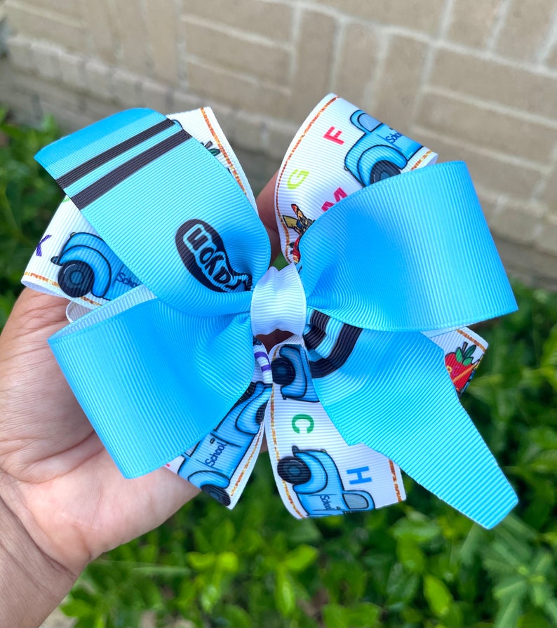 May include: A blue and white hair bow with a school bus design. The bus has the word "School" written on it and the letters "ABC" are printed on the bow.
