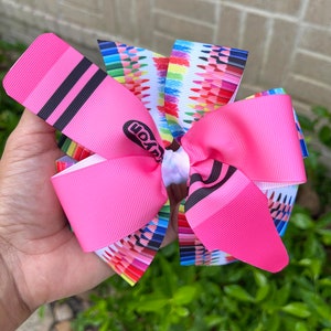 May include: A pink hair bow with a Crayola crayon print. The bow is made of two layers of pink ribbon with a white center. The crayon print features a variety of colors and is centered on the bow.
