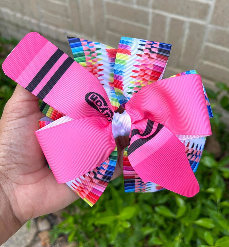 May include: A pink hair bow with a Crayola crayon print. The bow is made of two layers of pink ribbon with a white center. The crayon print features a variety of colors and is centered on the bow.