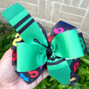 May include: Green and black hair bow with a colorful number and school supply print. The bow is tied with a black ribbon.