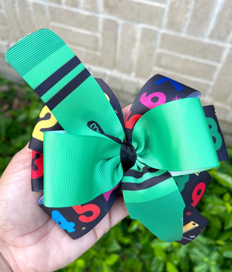 May include: Green and black hair bow with a colorful number and school supply print. The bow is tied with a black ribbon.