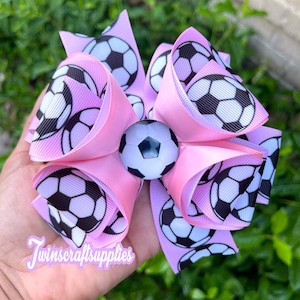 May include: Pink hair bow with a soccer ball design. The bow has a large, white soccer ball in the center. The text "Twincraftsupplies" is visible in the bottom left corner of the image.