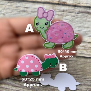 May include: Two pink and green turtle shaped wooden cutouts with white polka dots. The larger cutout is 50 by 40 mm and the smaller cutout is 50 by 25 mm. Both cutouts have a pink bow on top of their heads.
