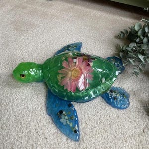 May include: A sea turtle figurine crafted from translucent resin. The shell features a large, pressed pink flower at the center, while the flippers and body contain embedded small, multi-colored stones and pebbles in shades of blue and green.