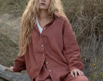 May include: A rust-colored linen shirt and pants set. The button-down shirt is paired with matching wide-leg pants. The outfit is worn with a white undershirt. The model is sitting on a log in a natural outdoor setting.