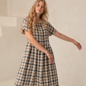 May include: A woman wearing a midi-length, short-sleeved dress with a plaid pattern in shades of beige, navy, and brown. The dress has a gathered waist and is paired with brown sandals. The background is a neutral-toned room with a staircase.