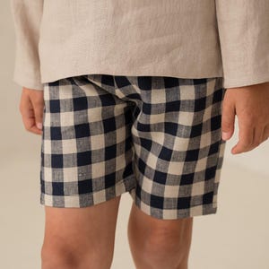 May include: A beige long-sleeve shirt paired with navy and white checkered shorts. The outfit is made of a soft, lightweight fabric, perfect for warm weather. The shorts feature a classic gingham pattern.